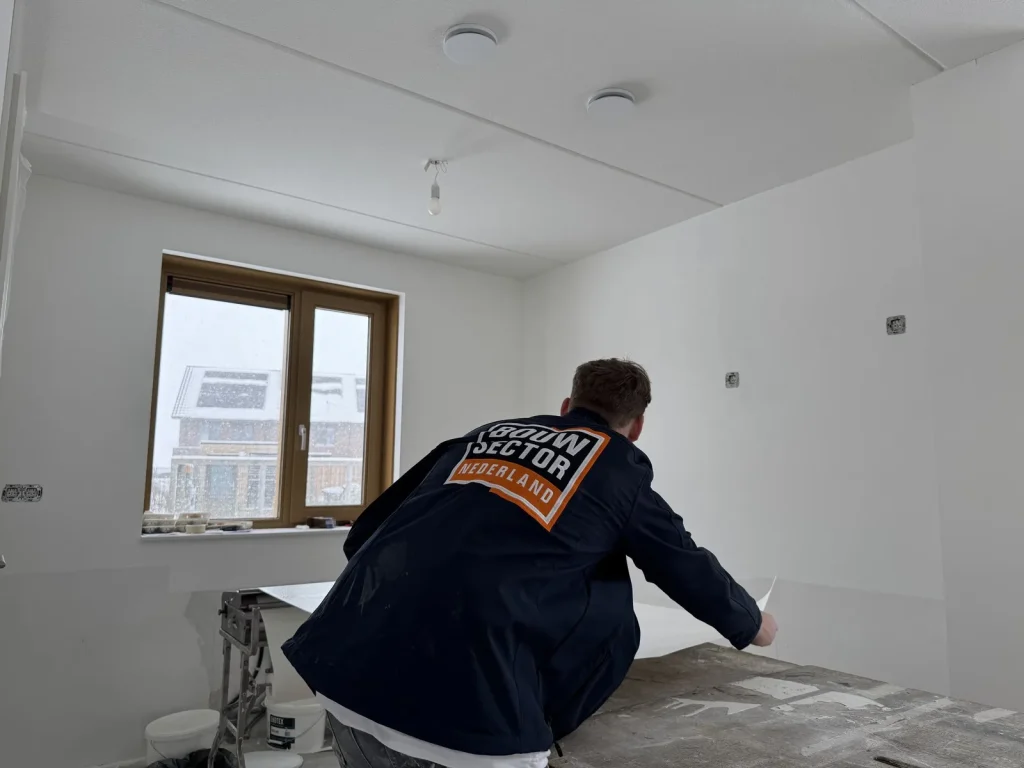 Behanger in bedrijfsjas pakt een baan vlies van de plaktafel in een nieuwbouwkamer met sneeuw buiten